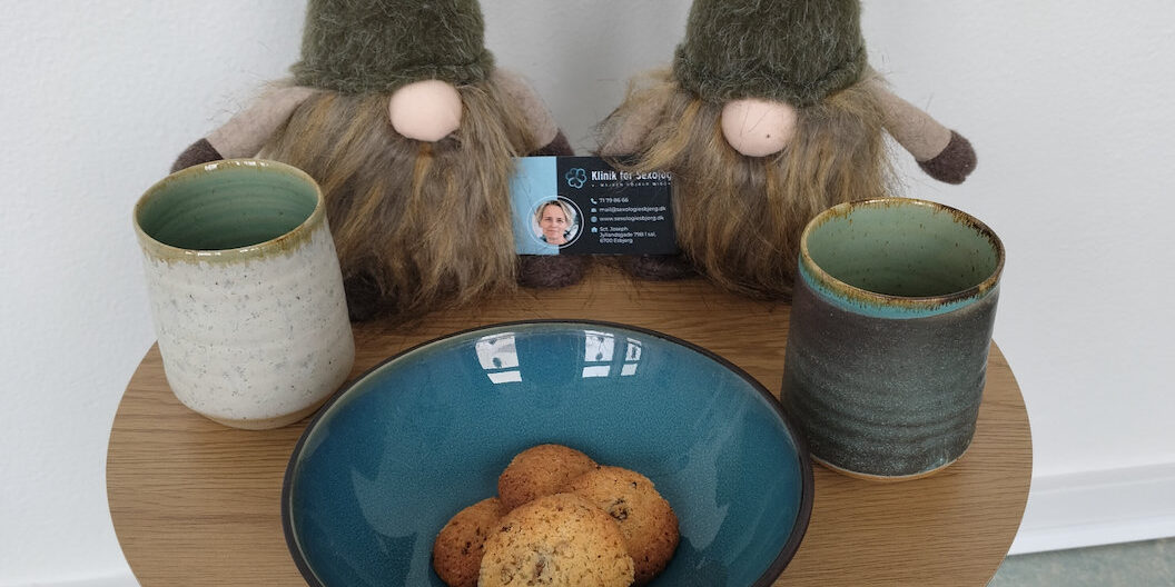 To jule-nisser med huerne trukket ned over øjnene står på et cafébord med to krus, småkager og visitkort fra Klinik for Sexologi – en påmindelse om nærvær og kærlighed i juletiden.