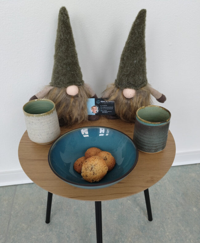 To jule-nisser med huerne trukket ned over øjnene står på et cafébord med to krus, småkager og visitkort fra Klinik for Sexologi – en påmindelse om nærvær og kærlighed i juletiden.