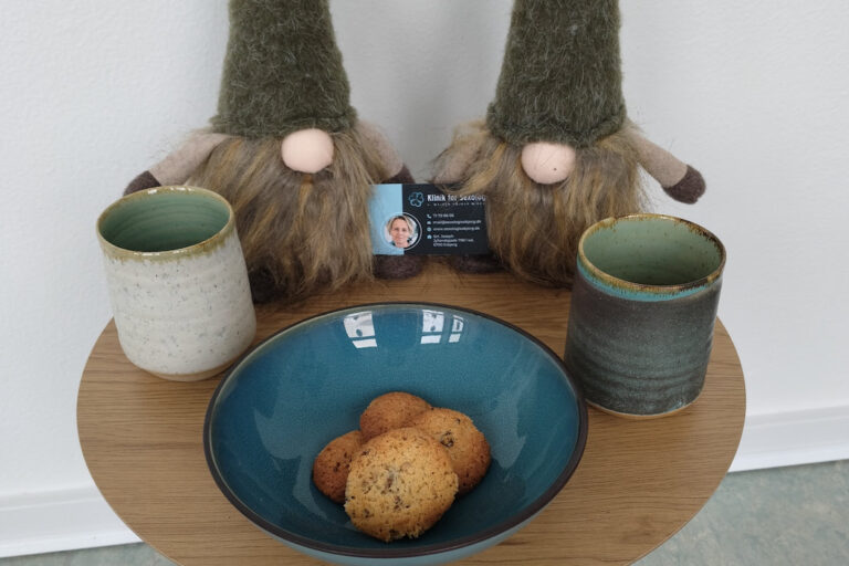 To jule-nisser med huerne trukket ned over øjnene står på et cafébord med to krus, småkager og visitkort fra Klinik for Sexologi – en påmindelse om nærvær og kærlighed i juletiden.