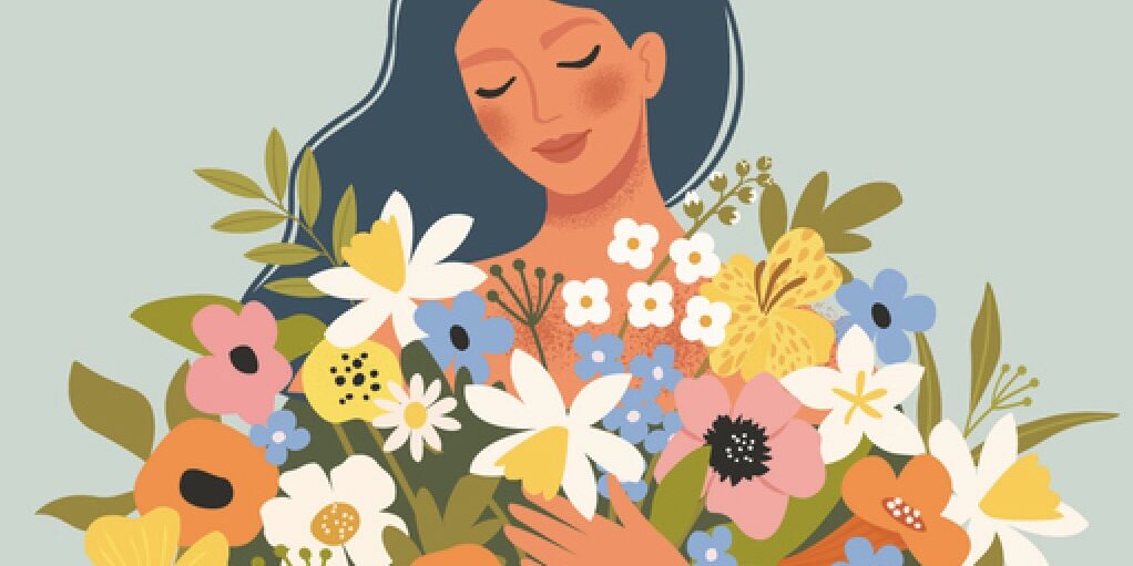 Smilende kvinde med lukkede øjne og favnen fuld af farverige blomster – et billede på styrke, eftertænksomhed og forandring i overgangsalderen.